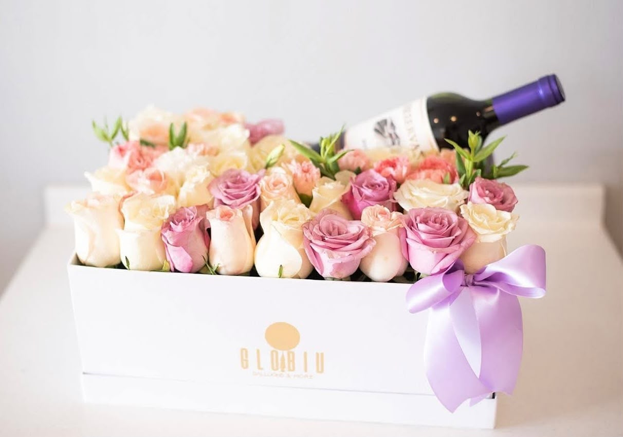 Caja floral con rosas en tonos crema y rosa, botella y lazo lila